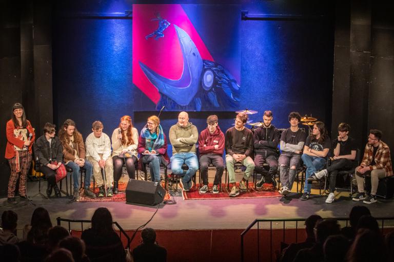 A photograph of teenage and adults artists sitting on a stage. A large artwork of a massive crow swallowing a knight is hung in the background