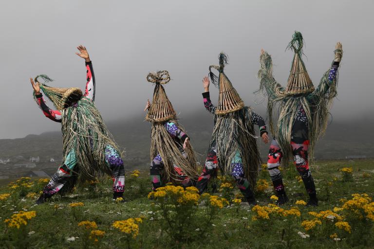 A photograph of 4 dancers wearing strawboy costume standing on a grassy area with heavy fog in the background
