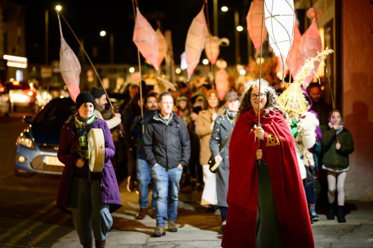 A photograph of a lantern parade at night. People are dressed in costume, holding lanterns and instruments while walking through a town