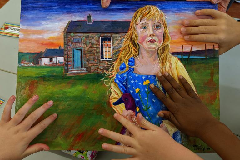 A photograph of childrens hands holding a painting of a blonde girl standing in front of a stone cottage