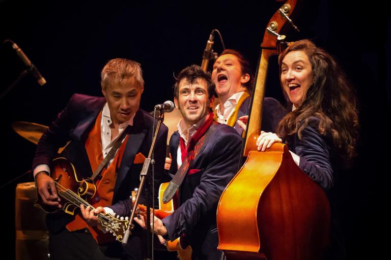 A photograph of four performers on stage singing into microphones and playing guitars and double bass