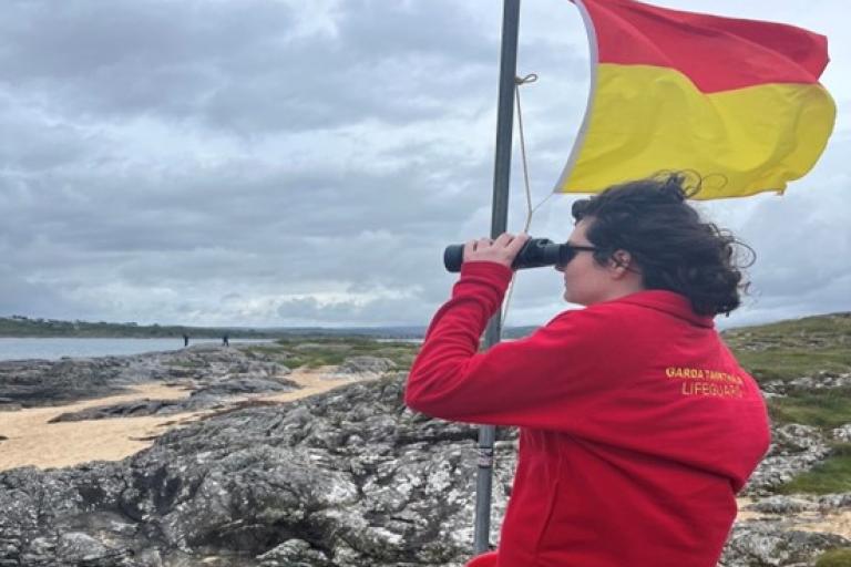 Lifeguard in red jacket with binoculars stands on rocks near a beach, beside a red-yellow flag. Cloudy sky and distant beachgoers in the background.