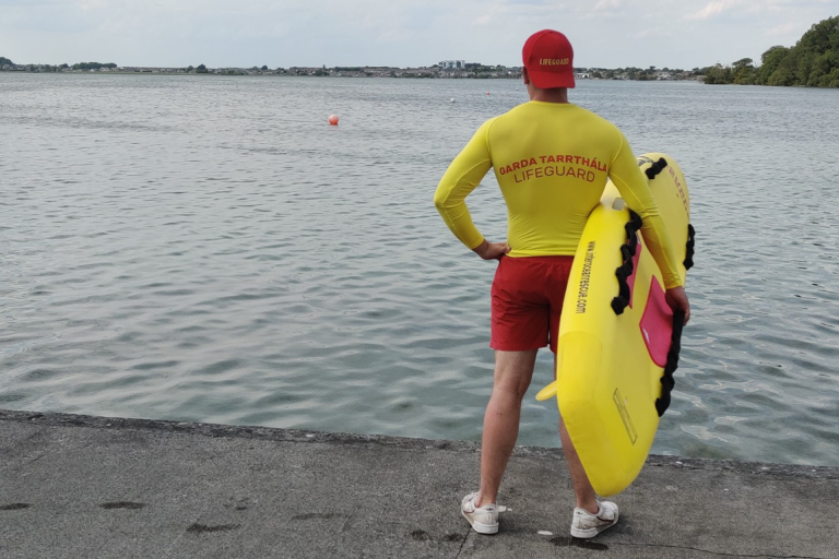 Lifeguard in red and yellow uniform holding a rescue board by calm water, with buoys and trees in the background.