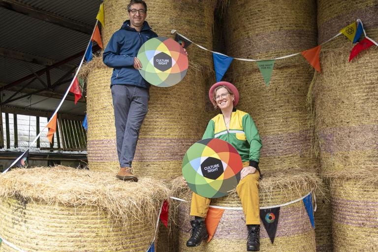 a man and woman sitting on hay bales holding colourful Culture Night signs