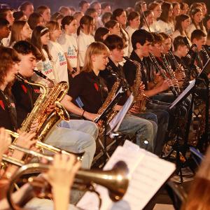 A photograph of a concert. Teenagers are playing brass and wind instruments seated with a children's choir standing behind