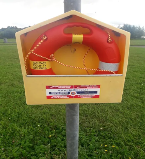 A red ring buoy with a rope is mounted inside a yellow box labeled "Galway County Council," with emergency contact info and a website below, set against a grassy background with trees.